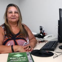 Mulher branca, cabelos loiros longos, com blusa marron em ambiente de escritório.