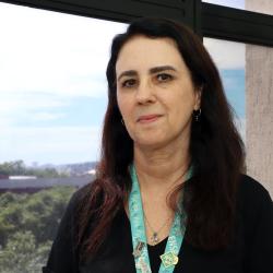 Mulher branca, com cabelos escuros longos, blusa preta em ambiente de escritório.