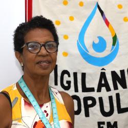 mulher da raça negra, cabelos escuros curtos, blusa amarela em frente a banner de vigilancia popular.