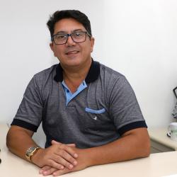 Homem Branco, com óculos de grau, camiseta polo cinza, cabelo curto preto em escritório.