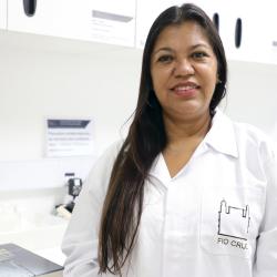 Mulher de cabelo escuros longos, jaleco branco da Fiocruz e dentro do laboratório.