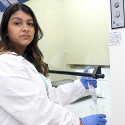 Mulher branca, cabelos escuros longos, com jaleco branco em ambiente de laboratório.
