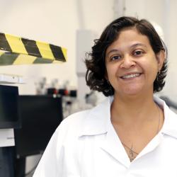 Mulher de cabelos escuros e curtos, com jaleco branco em atividade no laboratório.