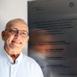 Homem branco com camisa branca e óculos de grau, próximo da placa de homenagem ao dssa..