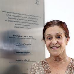 Mulher branca com cabelo preso em frente a placa de homenagem ao departamento