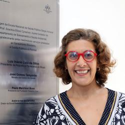 Mulher com blusa preto com detalhes branco, óculos de aro vermelho em frente a placa de homenagem ao dssa.