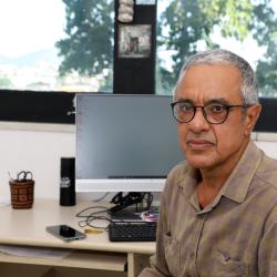 Homem com óculos de grau, camiseta marron claro em ambiente de escritório.