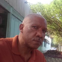 Homem com bigoe, camisa polo marron, em ambiente ao ar livre.