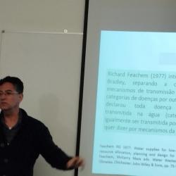 homem de cabelo preto e curto, camisa preta, lecionando em sala de aula.